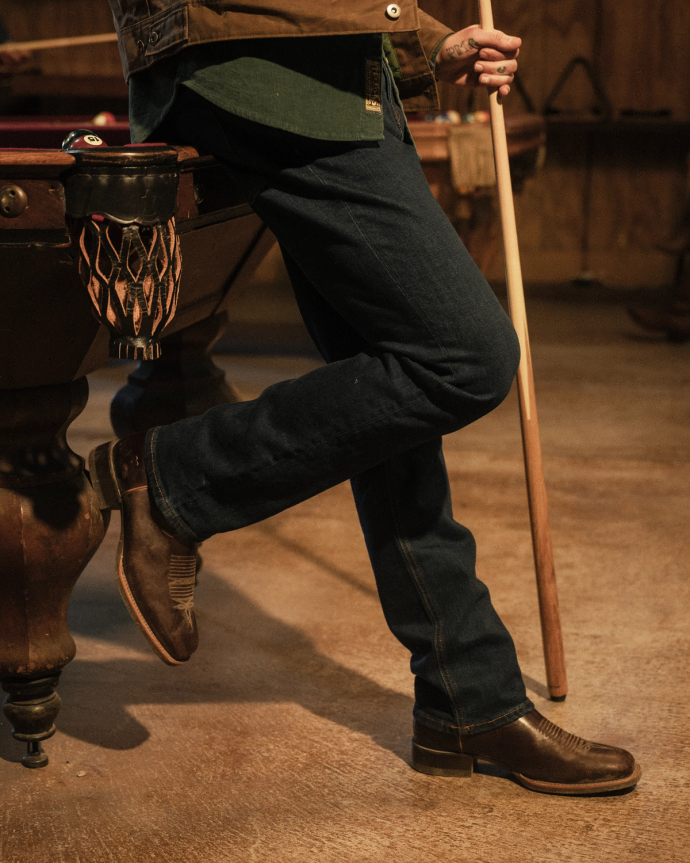 Person wearing brown boots and dark jeans leaning against a pool table while holding a cue stick.