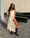 A woman in a sleeveless cream dress and black boots smiling on a sunny day outdoors.