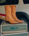 A pair of tan leather cowboy boots sit on top of two stacked green shoe boxes, with a white SUV in the background.