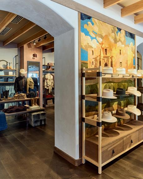 A store interior with shelves displaying hats and clothing. A large mural depicting a scene of the Alamo is on the wall. The ceiling features wooden beams.