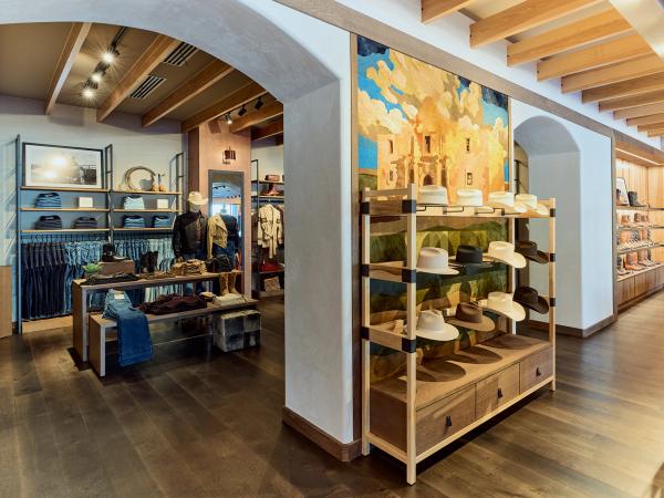 A store interior with shelves displaying hats and clothing. A large mural depicting a scene of the Alamo is on the wall. The ceiling features wooden beams.