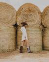 Person in a white dress and cowboy hat walking past large stacked hay bales, wearing brown boots.