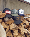 Four caps with various designs rest on a pile of cut firewood, set against a concrete wall background.