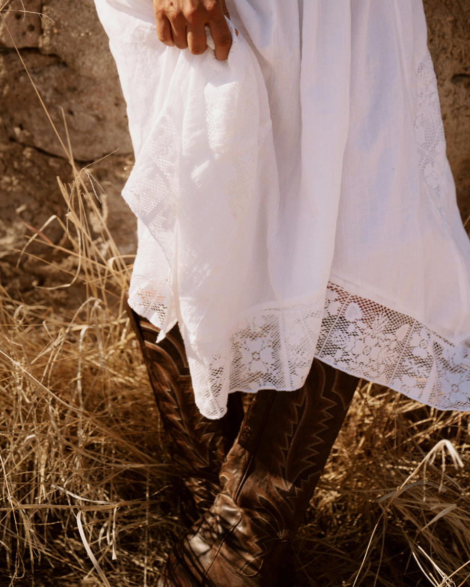 The Abby in Mocha Goat. Person in a white lace-trimmed dress holding the hem, standing in dry grass and wearing brown cowboy boots, with The Abby Goat grazing nearby.