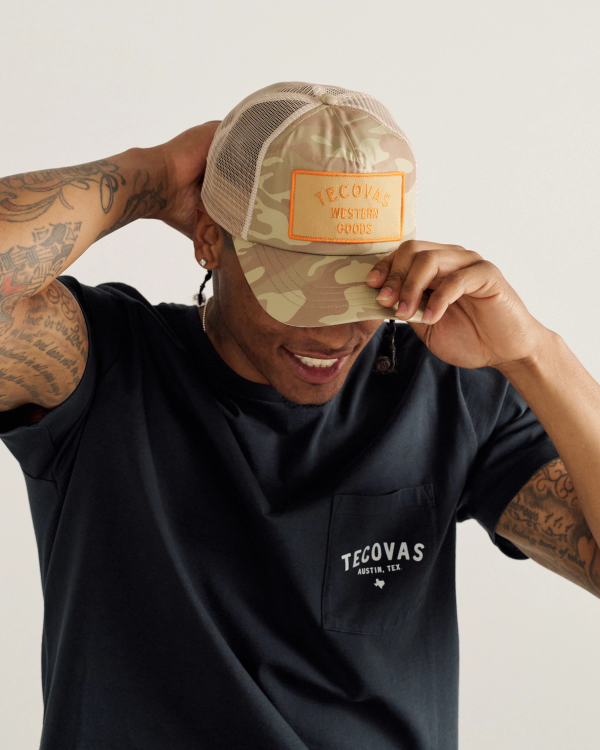 A person adjusts a camouflage Tecovas trucker hat while wearing a black Tecovas t-shirt.