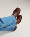 A person wearing blue jeans and brown leather cowboy boots with a textured pattern sits with legs crossed on a white background.