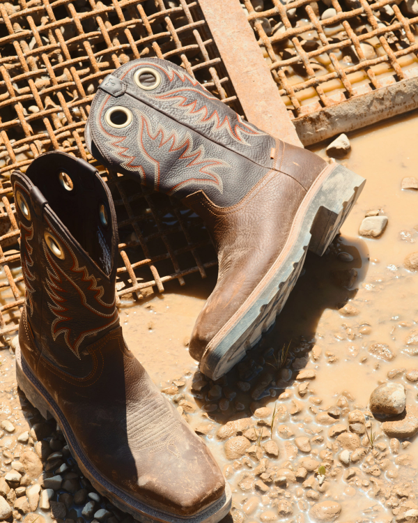 LongHaul Waterproof Square Composite Toe Work Boot in Tobacco / Black Cowhide. A pair of worn Tobacco / Black Cowhide cowboy boots with decorative stitching are placed on wet ground near a metal grate and muddy puddle.
