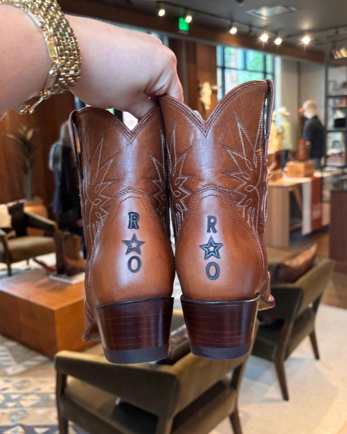 A hand holds up a pair of brown cowboy boots with white stitching and the letters "R" and "O" with stars underneath on the back of each boot.