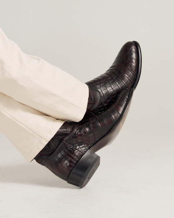 A person wearing light-colored pants and dark brown, crocodile-pattern leather boots with a low heel, resting their legs crossed.