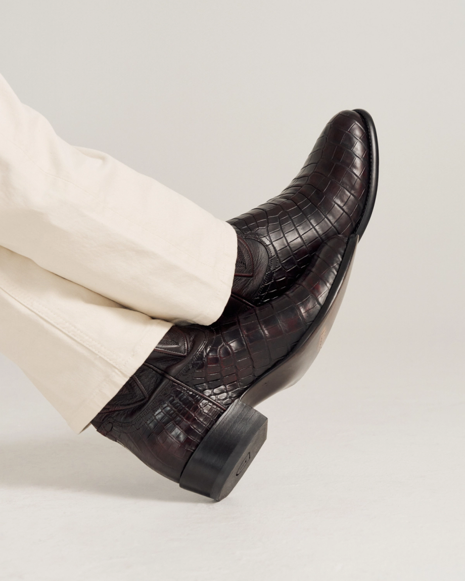 A person wearing light-colored pants and dark brown, crocodile-pattern leather boots with a low heel, resting their legs crossed.