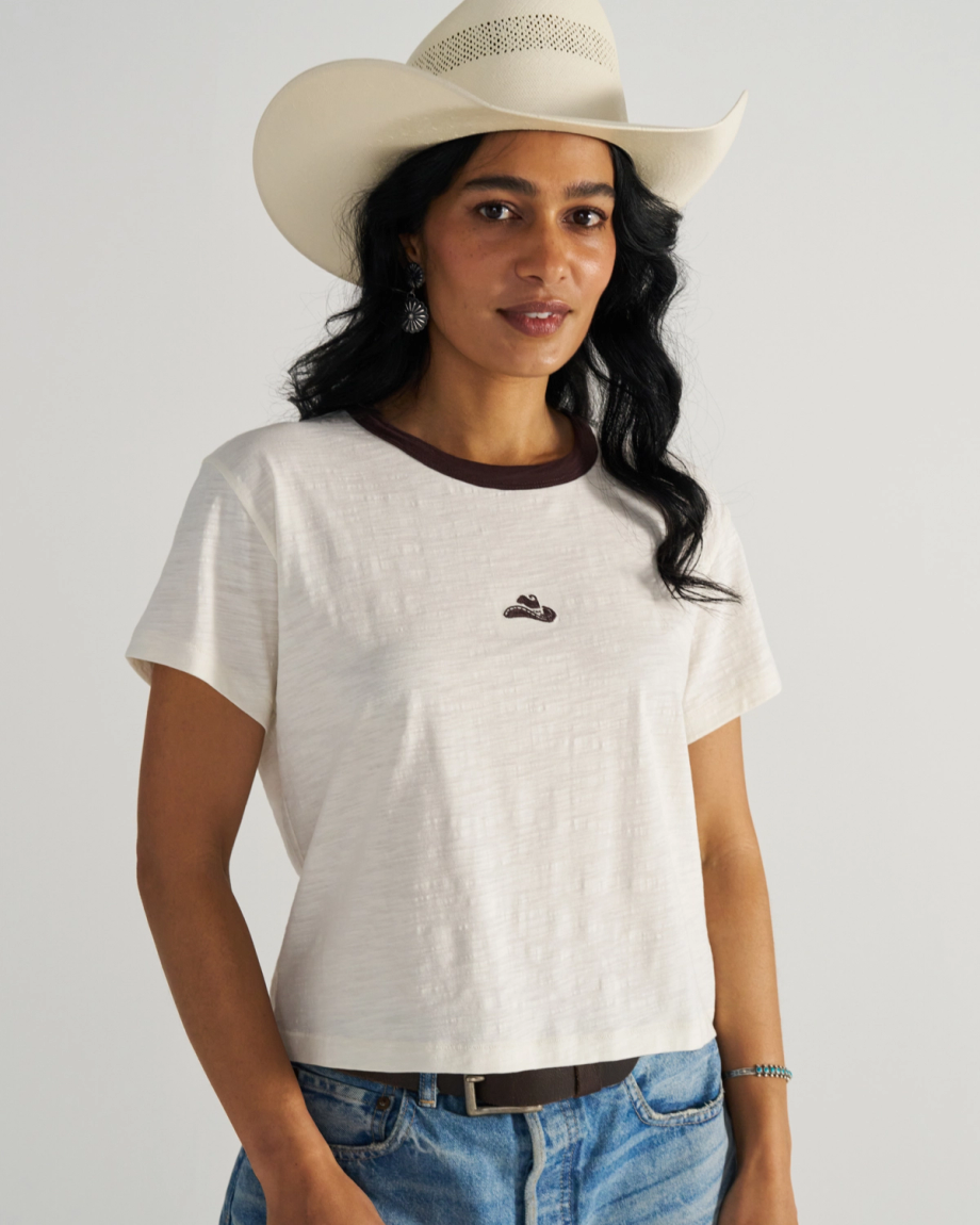 Woman in a white cowboy hat and white t-shirt with a small emblem, wearing blue jeans, stands against a neutral background.