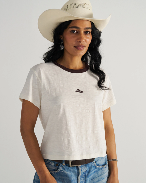 Woman in a white cowboy hat and white t-shirt with a small emblem, wearing blue jeans, stands against a neutral background.