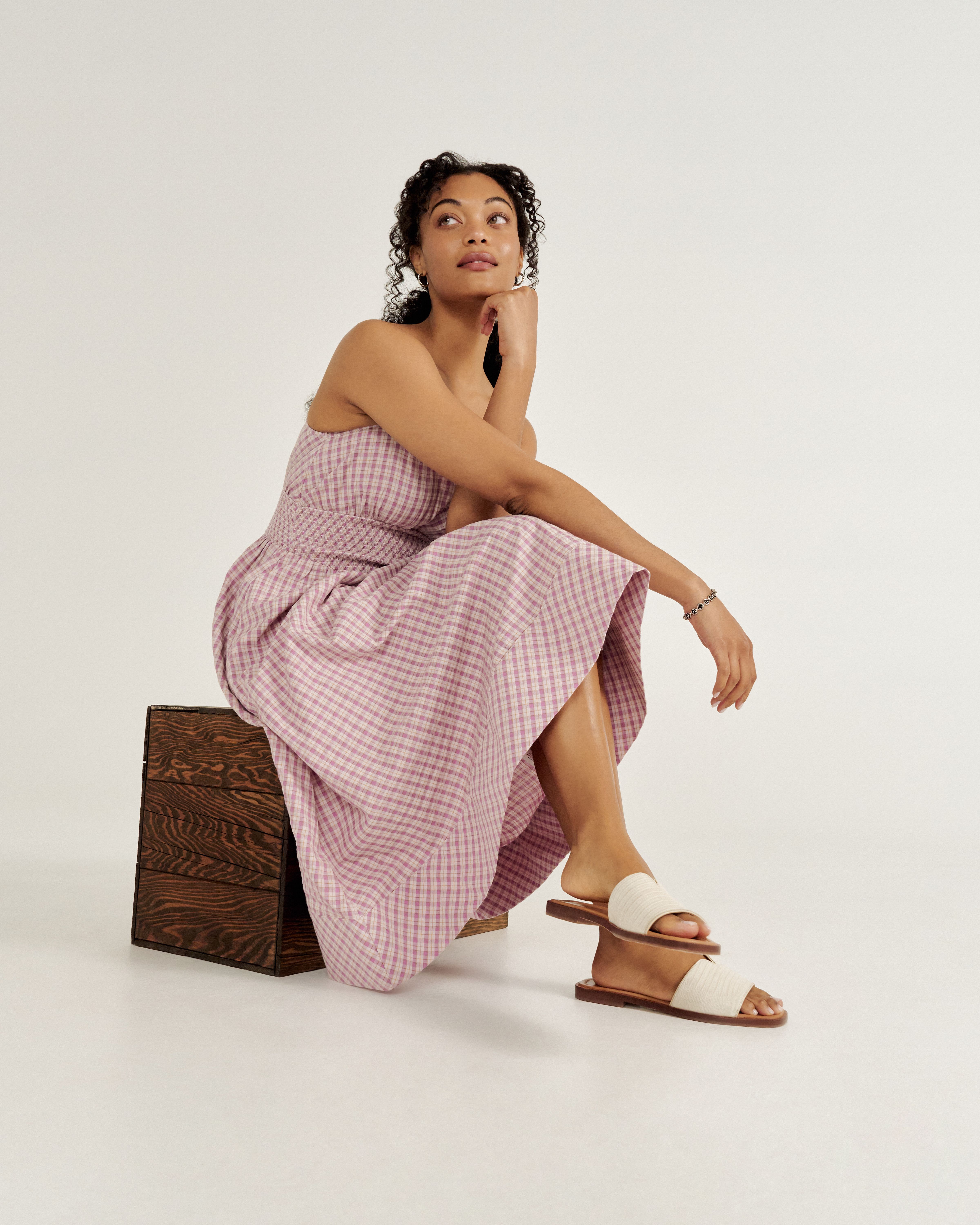 Person sitting on a wooden box, wearing a pink checkered dress and white sandals, looking upwards with one arm resting on their knee against a plain background.