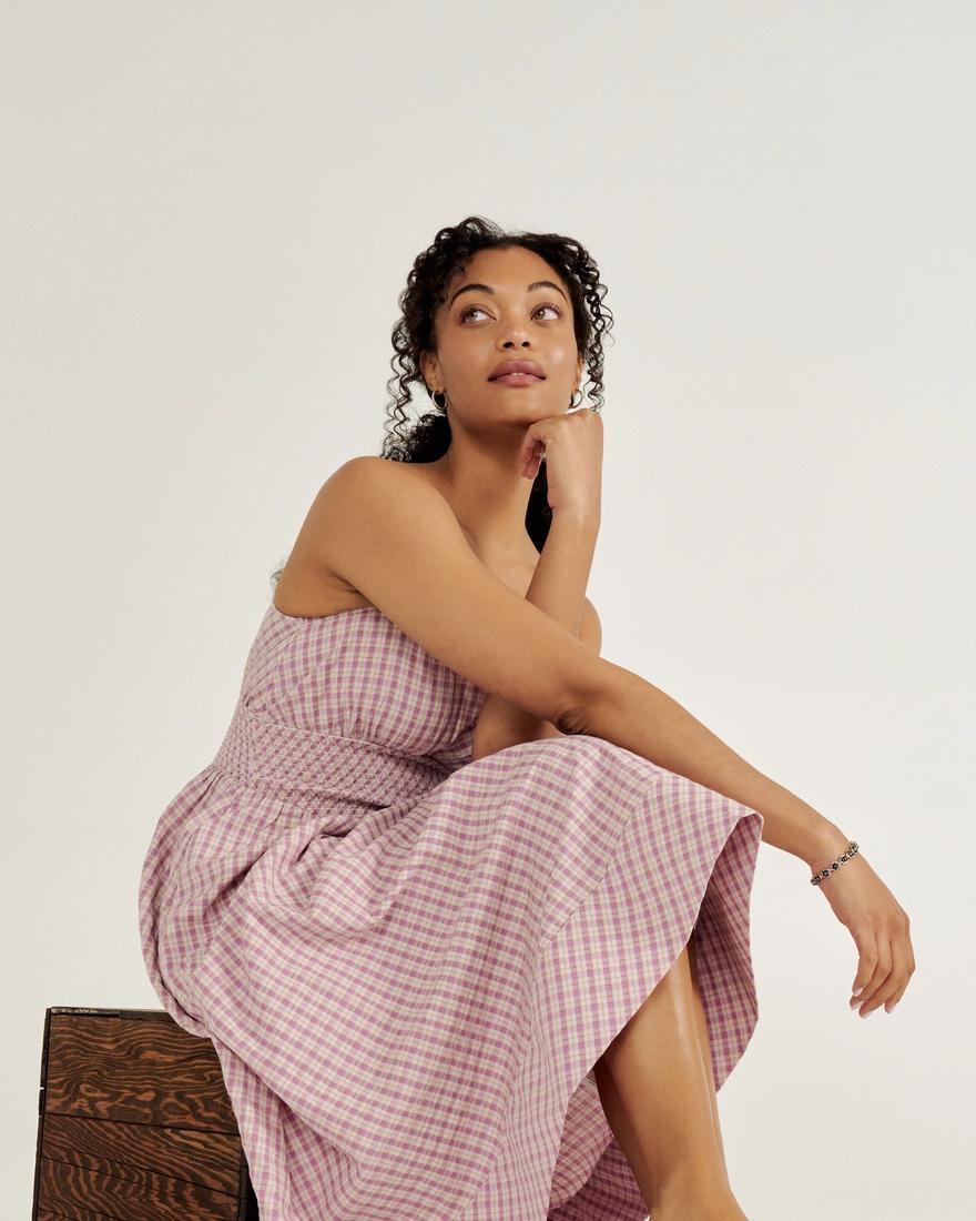 Person sitting on a wooden box, wearing a pink checkered dress and white sandals, looking upwards with one arm resting on their knee against a plain background.