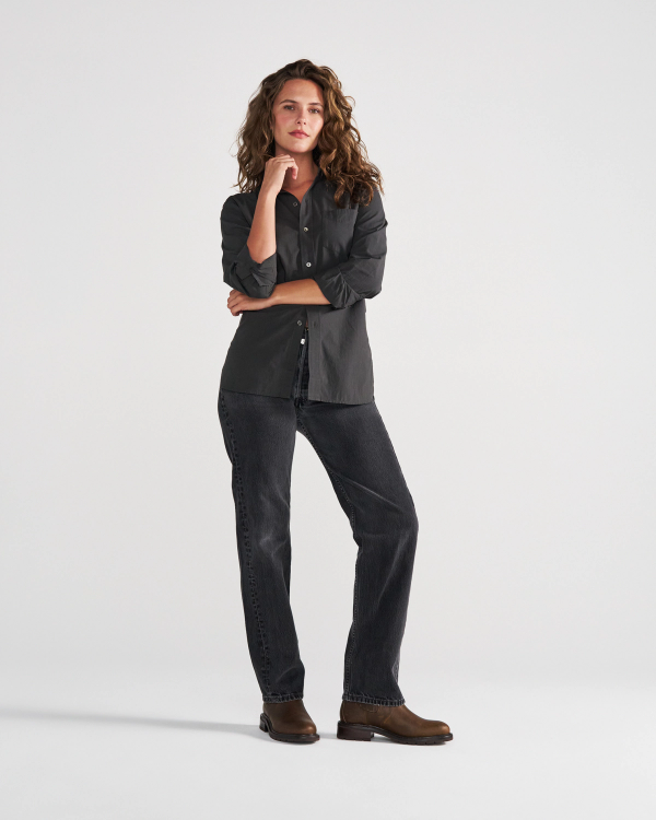 A person with long curly hair, wearing a dark button-up shirt, dark jeans, and brown boots, stands with one hand on their chin against a plain white background—evoking the relaxed yet stylish atmosphere of The Chelsea Cowhide cafe.