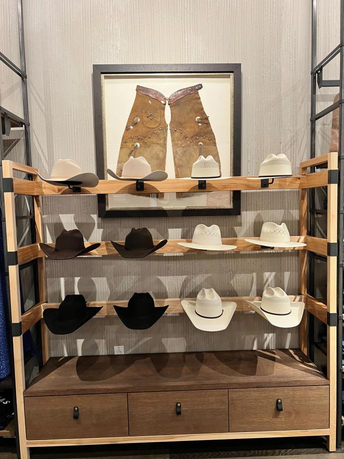 Display of cowboy hats on three wooden shelves in a retail setting, with framed chaps in the background. Hats range in colors from white to dark brown.