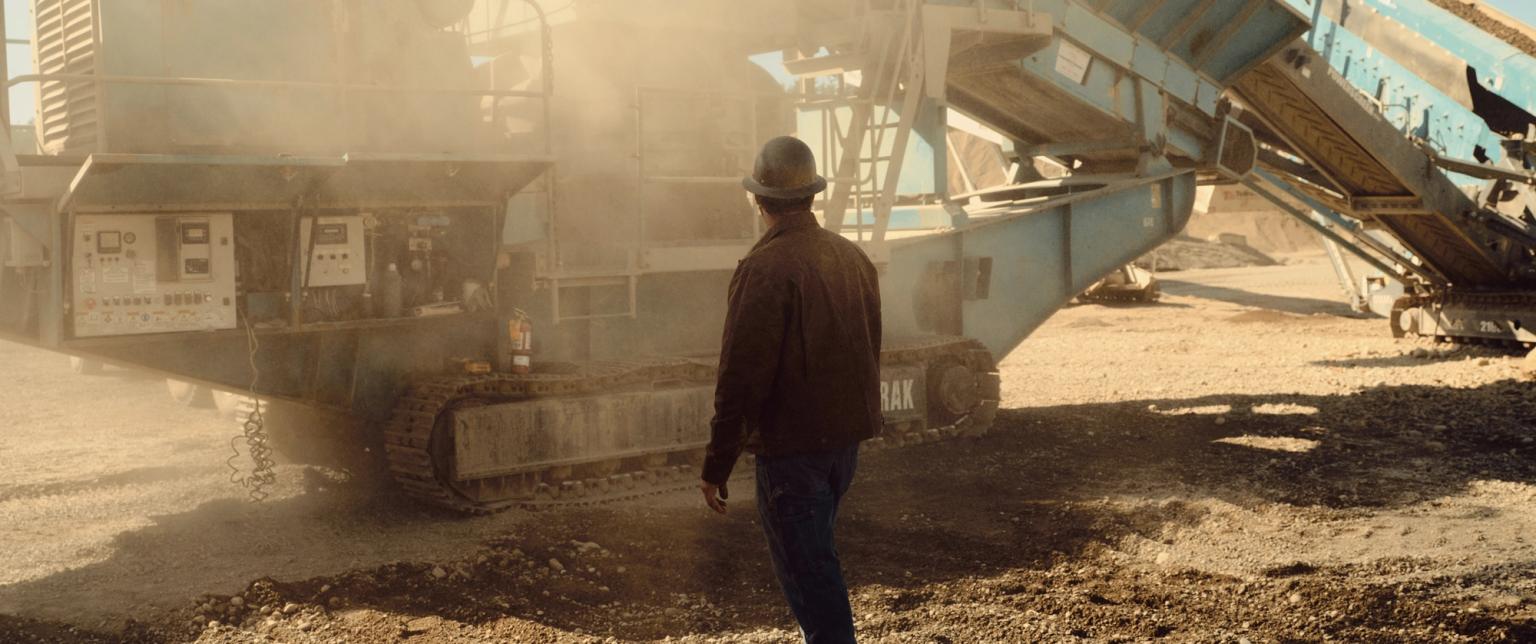 A worker in a hard hat and jacket stands near large industrial machinery on a dusty construction site.