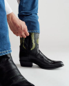 A person wearing blue jeans pulls on a black cowboy boot with green and black detail against a plain background.