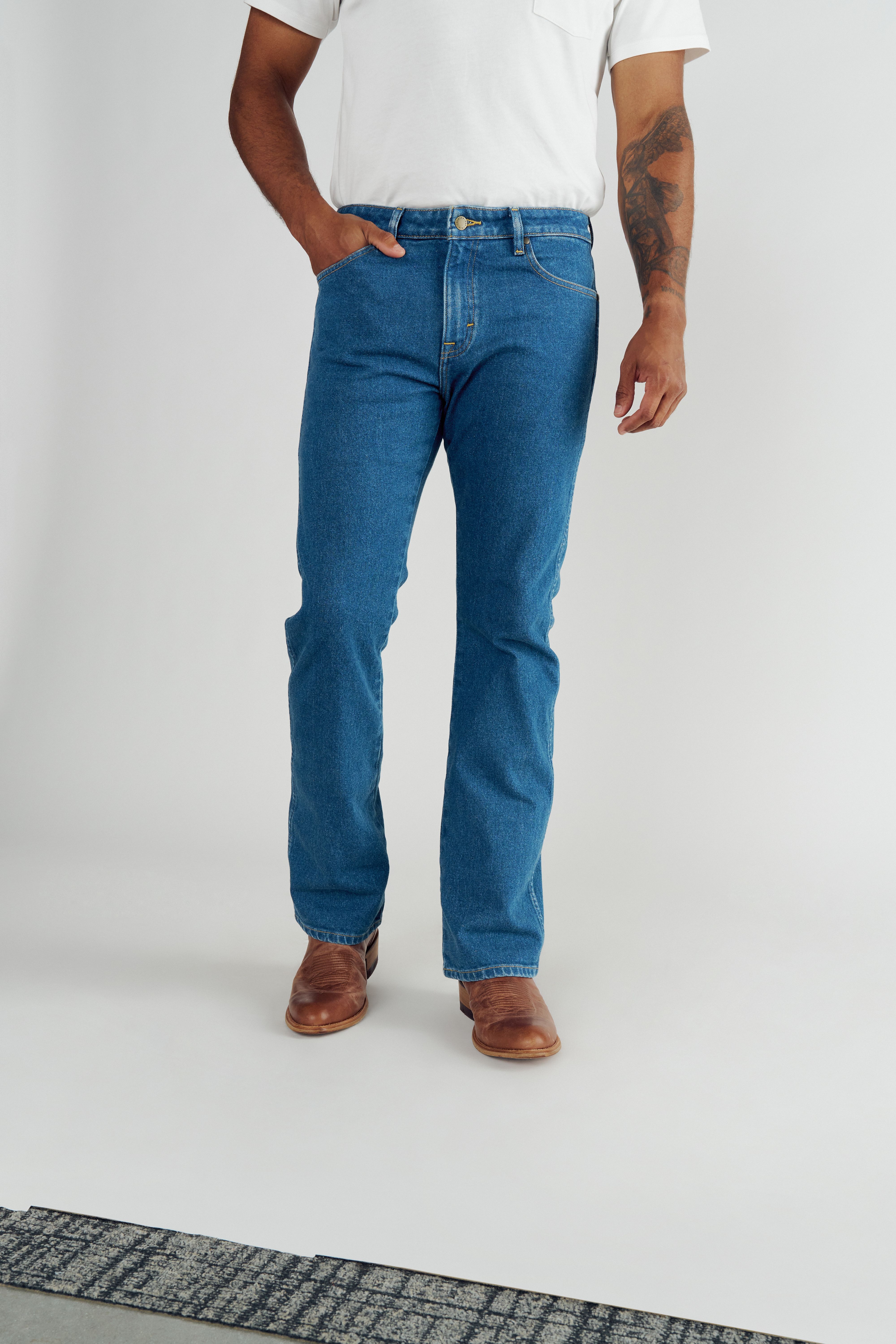 A person wearing a white t-shirt, blue jeans, and brown boots stands with one hand in their pocket against a plain background.