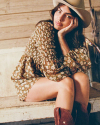 Person seated on a wooden bench, wearing a floral dress, hat, and boots, resting cheek on one hand and looking towards the camera.