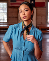 A woman in a blue striped dress and a patterned scarf tied around her neck stands in a room with historical flags in the background.