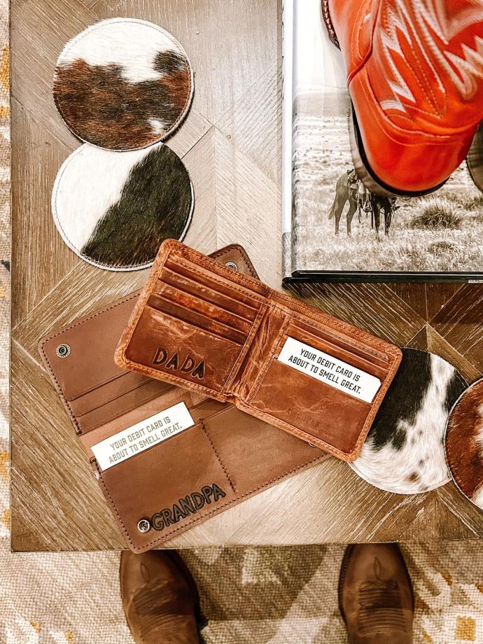 A brown leather wallet marked "DADA" with a slot for credit cards is lying open on a wooden table. Nearby is an engraved leather item labeled "GRANDPA" and a pair of boots.