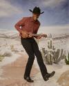 Man in cowboy hat, printed button up, black jeans and cowboy boots dancing with a mini guitar.