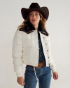 Woman wearing a black cowboy hat, white puffer jacket with dark collar, and denim shirt and jeans, standing against a plain background.