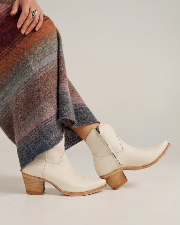 A person wearing cream-colored, mid-calf heeled cowboy boots and a multicolored, textured skirt standing on a light surface.