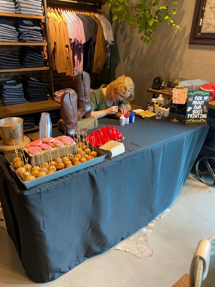 Person seated at a table with a displayed assortment of snacks, boots, and clothing. Sign in the background reads, "ASK ABOUT BOOT PAINTING.