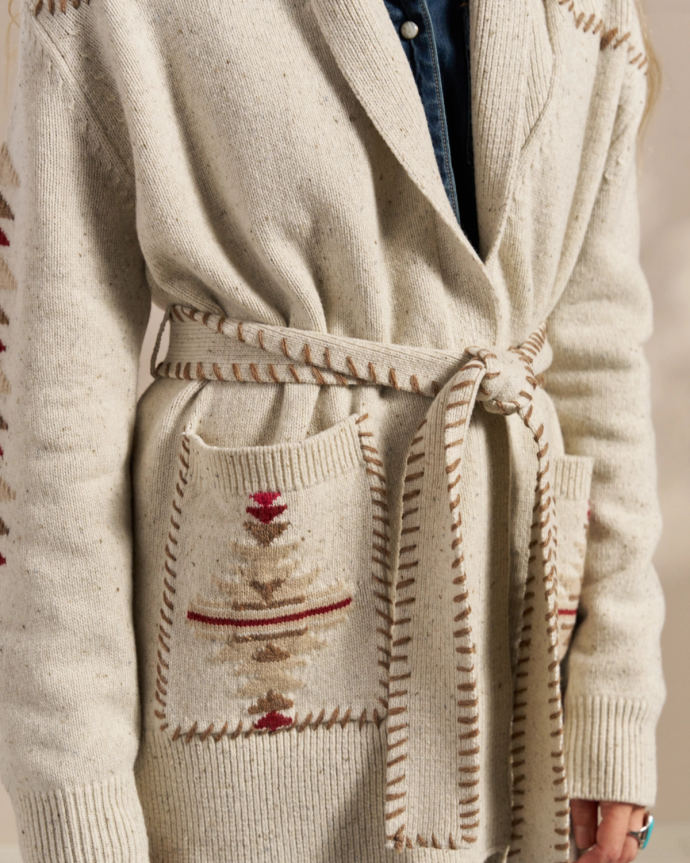 Close-up of a person wearing a beige knitted cardigan with a belt tied at the waist and patterned pockets. The cardigan has brown and red geometric designs.