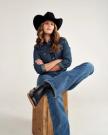 A person wearing a cowboy hat, denim shirt, and jeans sits on a wooden box with one leg crossed.