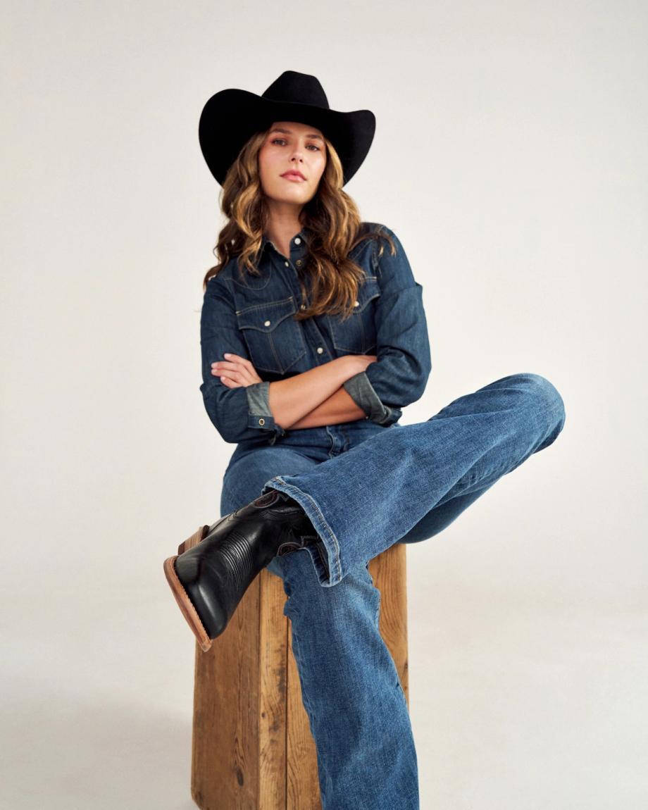 A person wearing a cowboy hat, denim shirt, and jeans sits on a wooden box with one leg crossed.