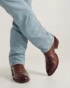 Person wearing light blue jeans and brown leather cowboy boots stands with legs crossed against a plain background.