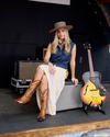 A woman in a bohemian style outfit, including a wide-brimmed hat and cowboy boots, sit beside an acoustic guitar.