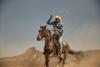 A cowboy in a denim jacket and straw hat swings a lasso while riding a galloping horse across the desert.