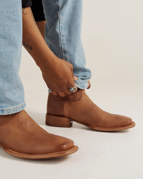A person wearing light blue jeans adjusts the cuff of their pant leg above a brown leather ankle boot.