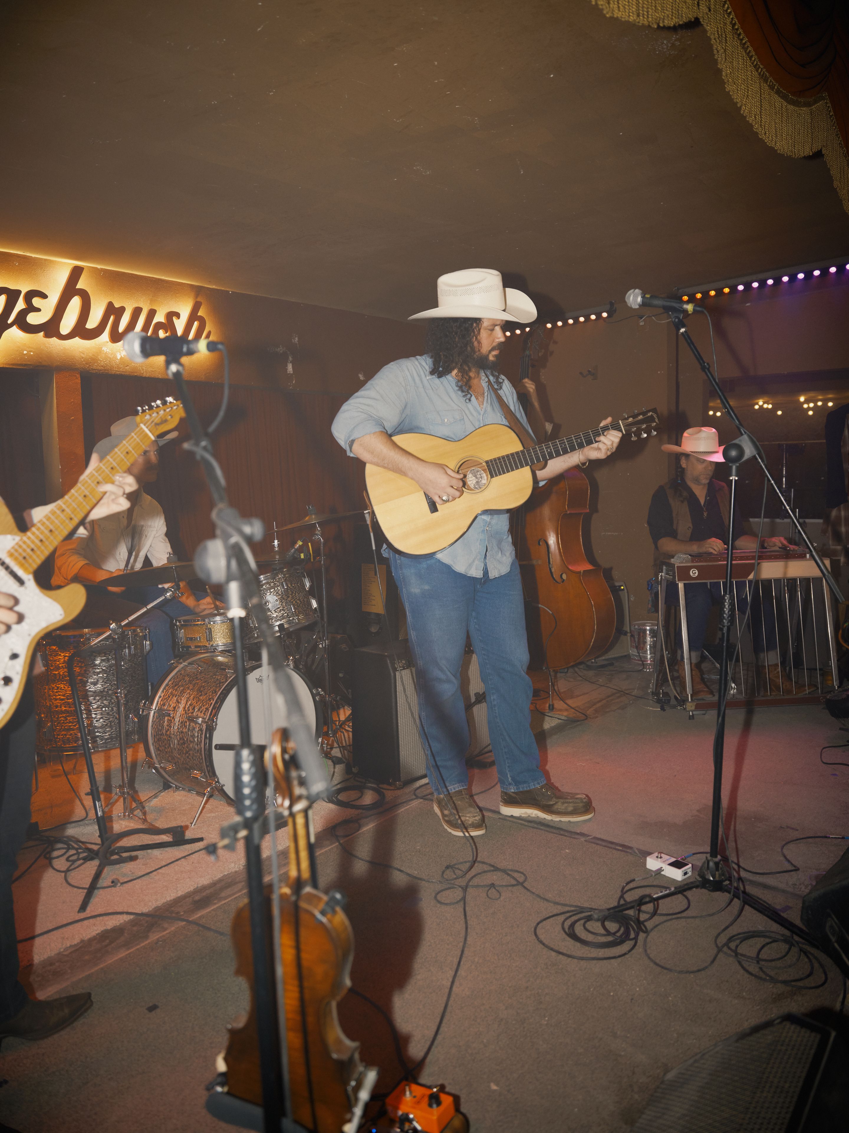 A musician in a hat plays an acoustic guitar on stage with a band, featuring drums and a steel guitar, under warm lighting.