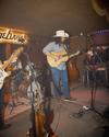 A musician in a hat plays an acoustic guitar on stage with a band, featuring drums and a steel guitar, under warm lighting.