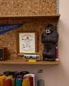 A shelf displays a framed illustration, a rubber gorilla figurine, a digital clock showing 12:08, and a blue pennant. Rolls of fabric and art supplies are visible on the lower shelf.