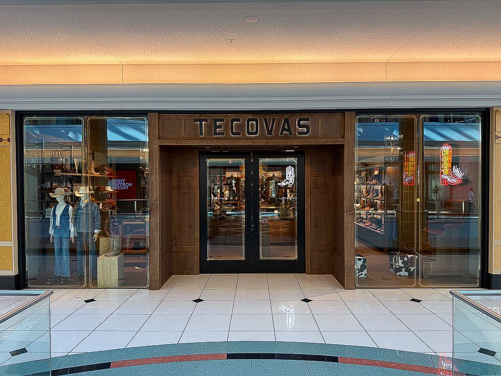Storefront of Tecovas with double glass doors, western boots, hats, and apparel displayed inside; tiled floor in front and glass railings on each side.