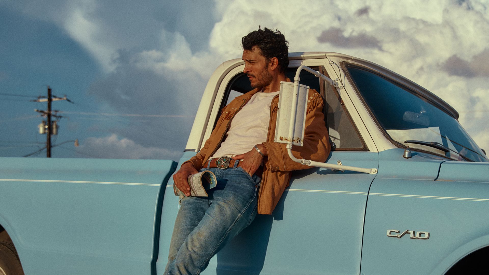 A man in a brown jacket and blue jeans leans against a light blue C10 pickup truck, holding a book, with clouds and power lines in the background.