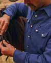 Man wearing a Tecovas blue pearl snap and cowboy hat adjusts the bridle on a horse.