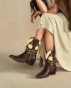 A seated woman in a light dress and brown jacket wears brown cowboy boots with yellow flower and green leaf designs.