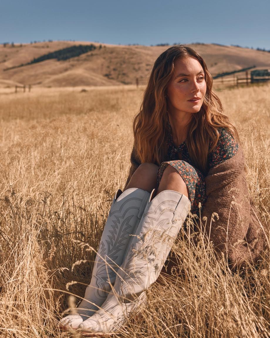 Woman sitting in a grassy field against a backdrop of hills, wearing a floral dress, brown shawl, and white boots.