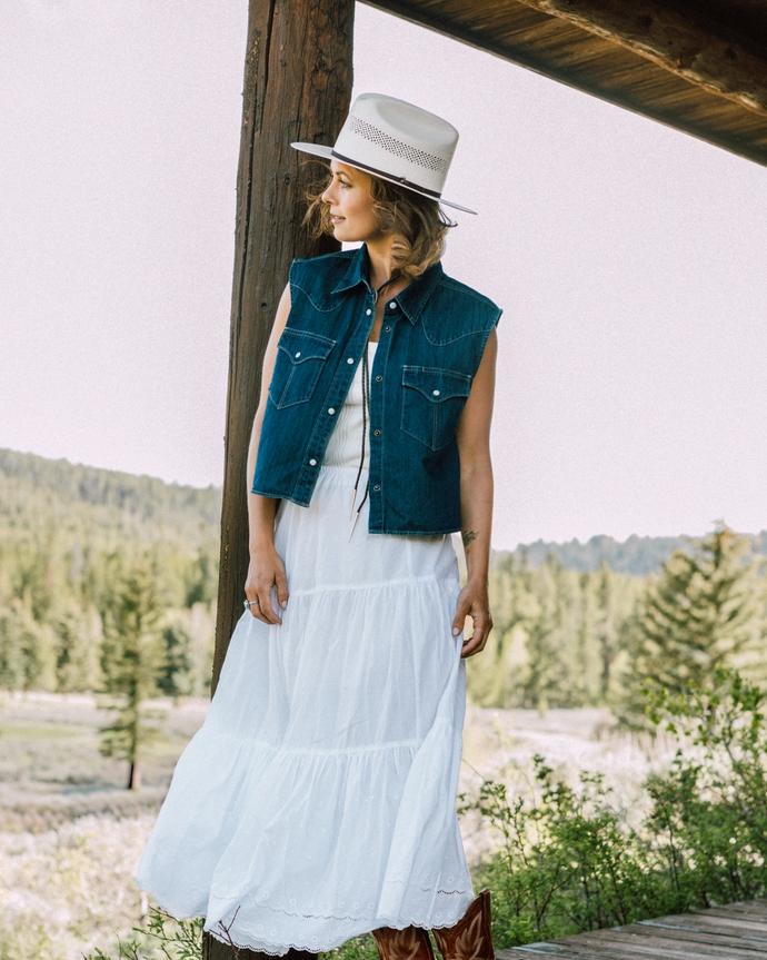 Woman standing on a porch wearing a denim vest and white skirt with a straw sun hat