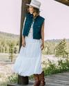 A woman wearing a white dress, denim vest, cowboy boots, and a hat stands on a wooden platform in a rural outdoor setting with trees and hills in the background.