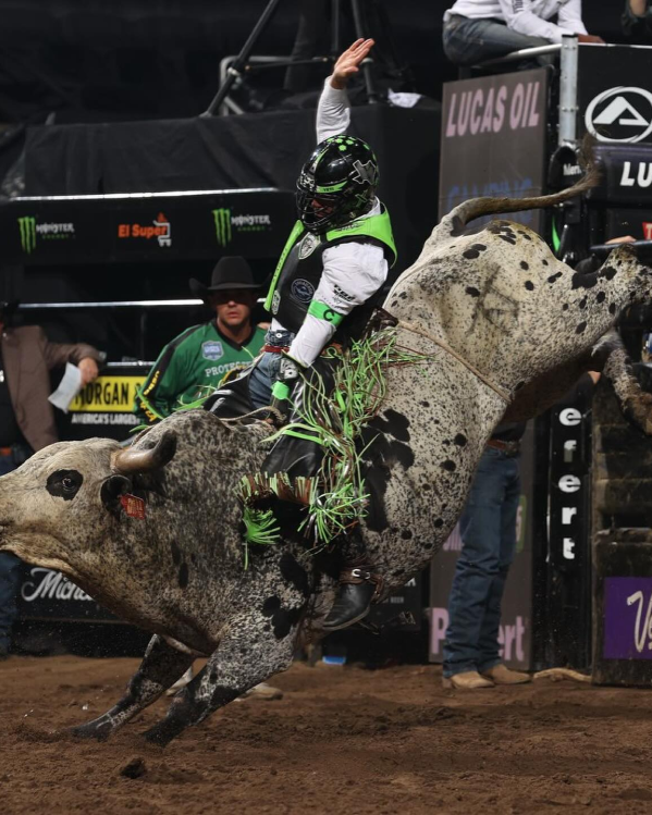 A rodeo participant rides a bucking bull in an arena, surrounded by spectators and event banners.
