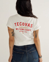 Woman wearing a white Tecovas t-shirt with red text that reads "Makers of Fine Western Goods, Austin, Tex," paired with blue jeans, standing sideways.