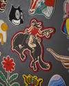 A collection of embroidered patches includes a cowboy on a bucking horse, a black and white mask, a floral pattern, and various animal designs on a textured dark gray background.